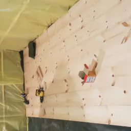 Wnętrze domu w trakcie budowy z widoczną izolacją w kolorze żółtym oraz drewnianą ścianą z zamontowanymi puszkami elektrycznymi i wkrętarkami.