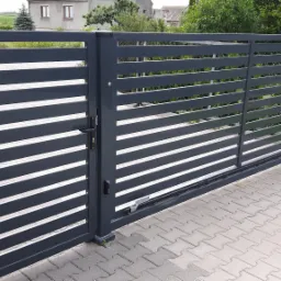 Nowoczesna, przesuwna brama wjazdowa z poziomymi panelami w kolorze grafitowym, zautomatyzowana, widoczny napęd i oświetlenie ostrzegawcze, kostka brukowa na podjeździe.