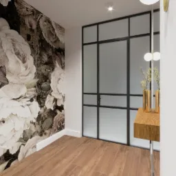 Wnętrze przedpokoju z fototapetą w kwiaty, drewnianą podłogą i przeszklonymi drzwiami w czarnej ramie, złote wazony na konsoli.