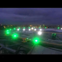 Wieczorny widok na oświetlony pas startowy dla helikopterów z zielonymi i białymi światłami nawigacyjnymi, z widoczną panoramą miasta w tle pod ciemniejącym niebem.