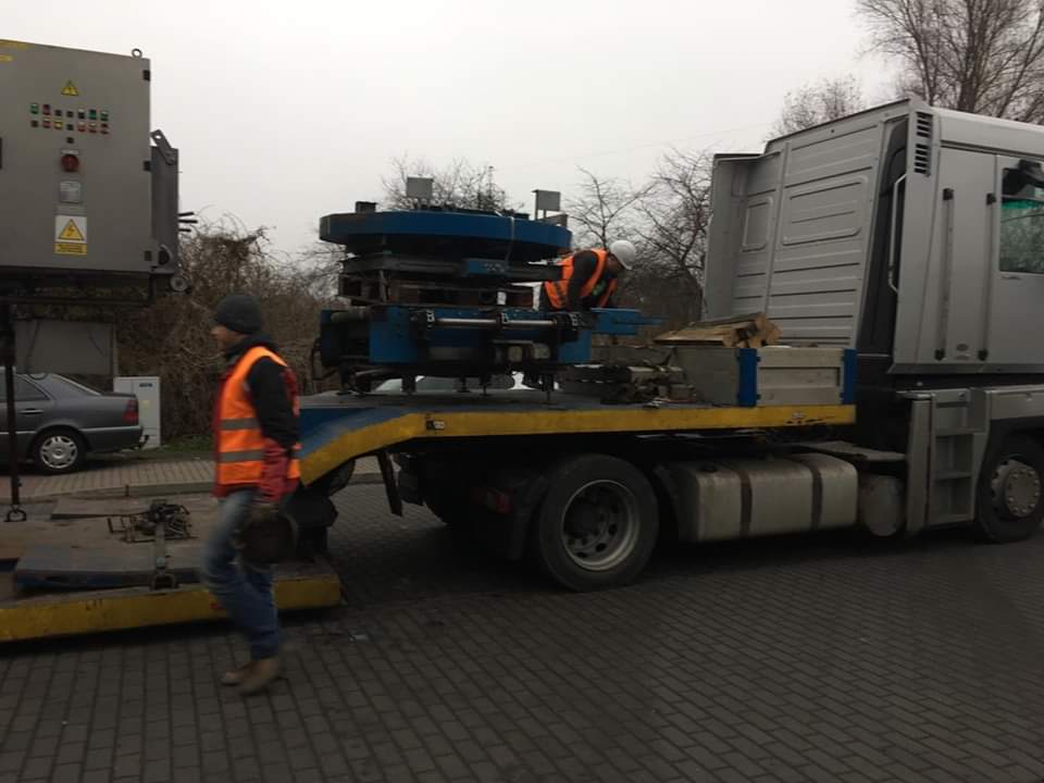 Transport transformatora na niskopodwoziowej platformie ciężarówki, dwóch pracowników w kamizelkach odblaskowych zabezpiecza ładunek.