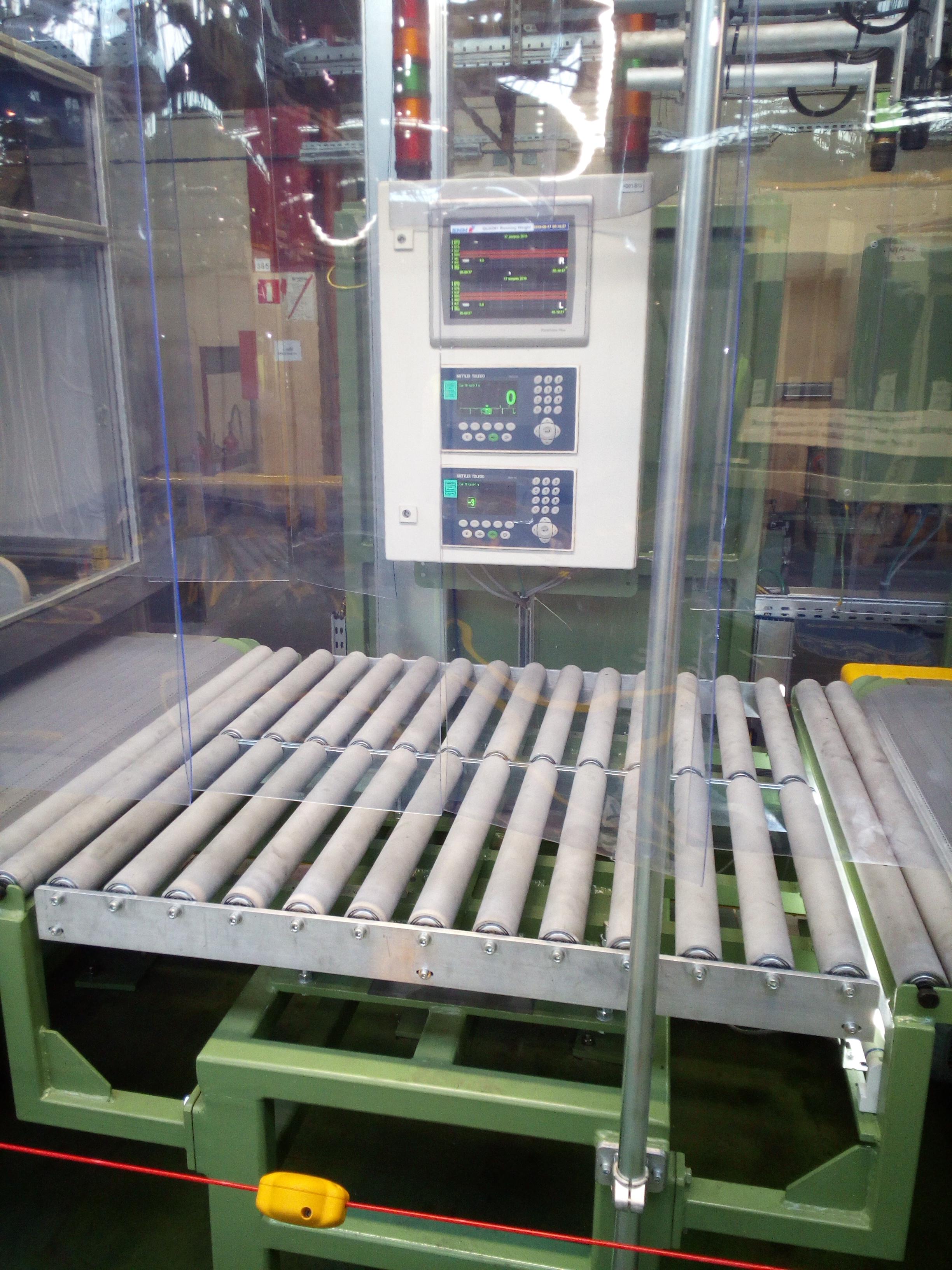 Przemysłowy system transportowy z rolkami, zabezpieczony przezroczystą osłoną, z panelem sterowania Mettler Toledo i czujnikiem laserowym bezpieczeństwa.