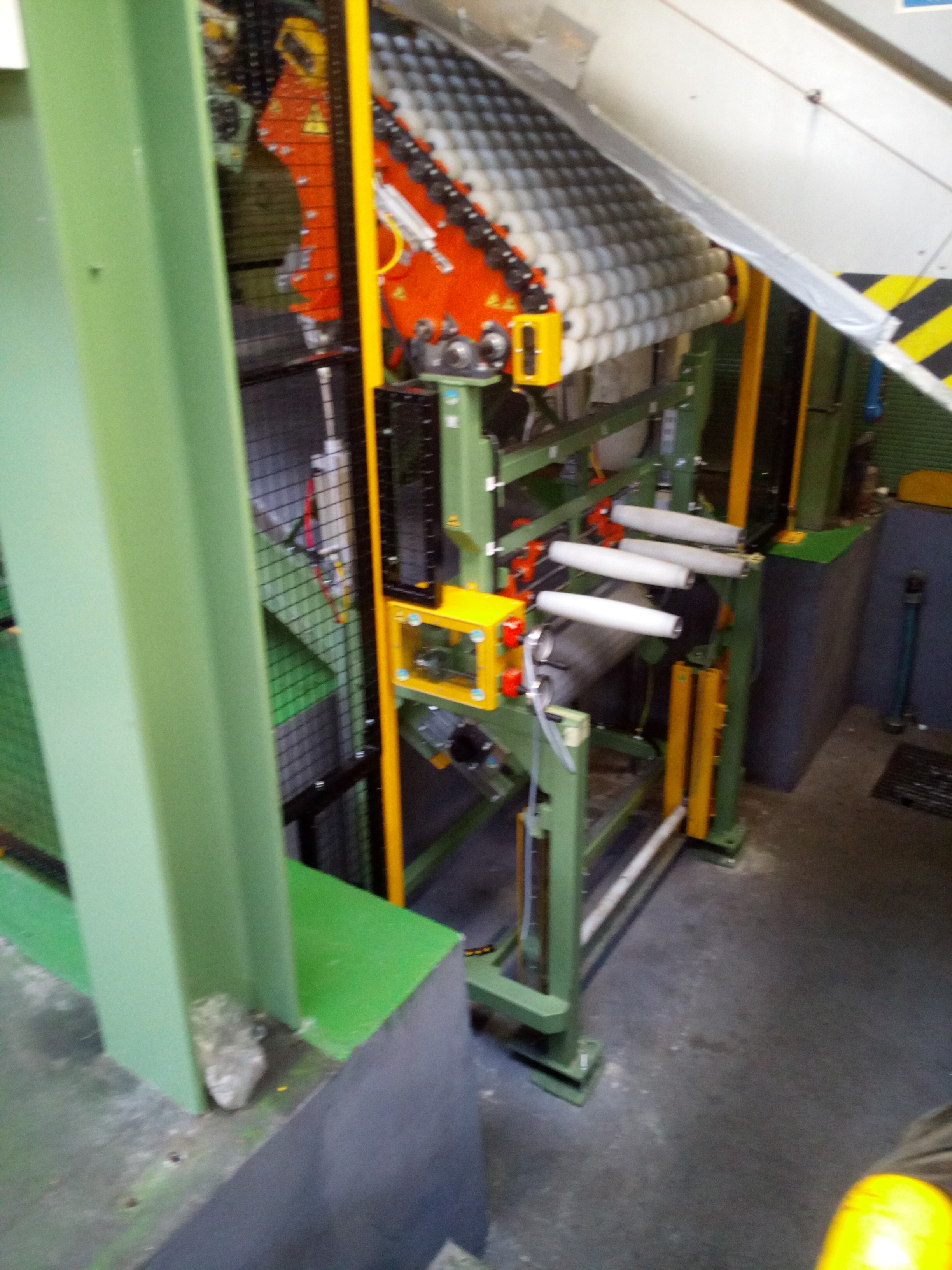Widok na fragment linii produkcyjnej z rolkami transportowymi, elementami automatyki i metalową konstrukcją w odcieniach zieleni i żółci.