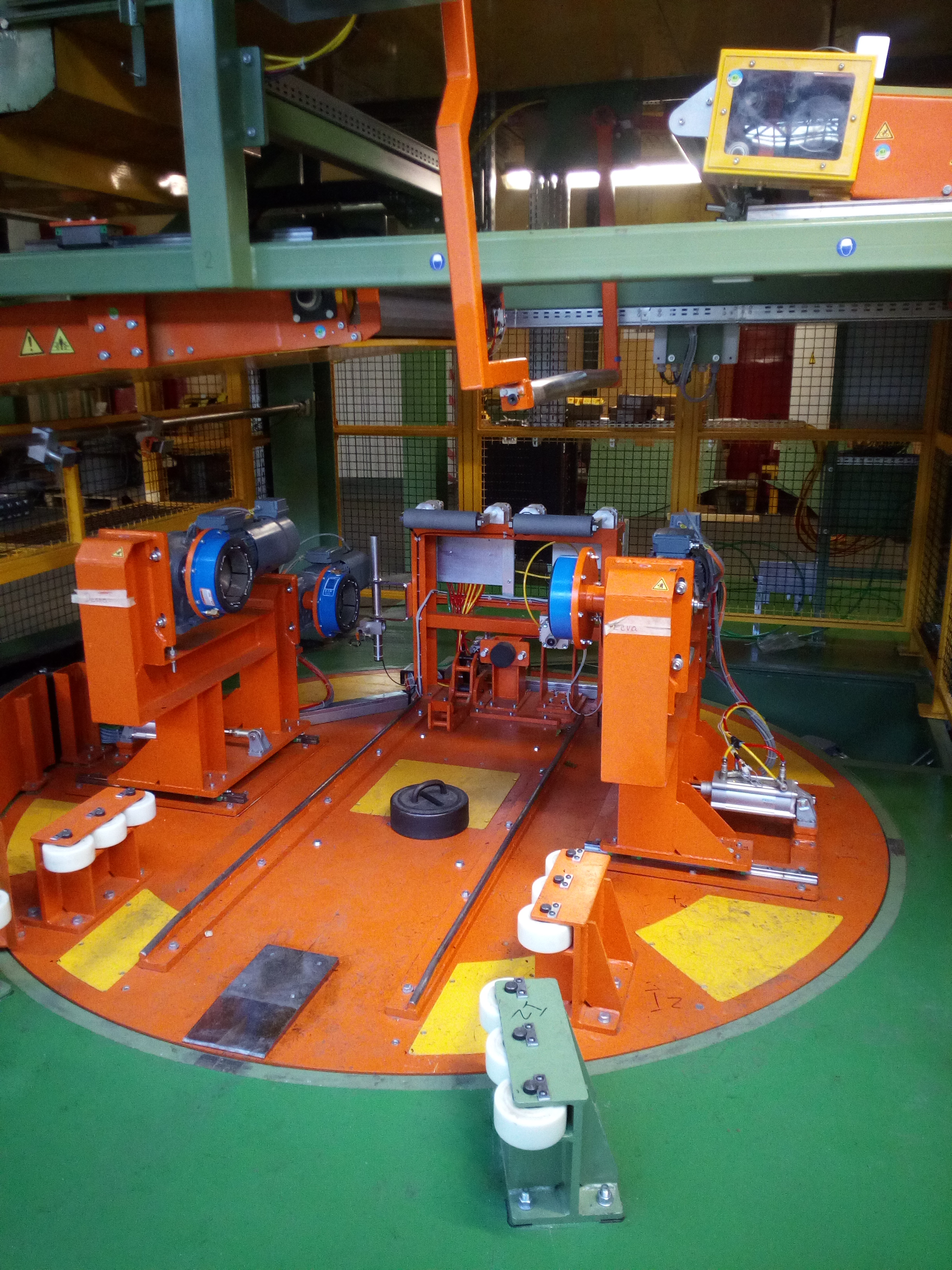 Detale automatycznej linii produkcyjnej z pomarańczowymi elementami konstrukcyjnymi, widoczne silniki elektryczne z niebieskimi obudowami, żółte przewody i metalowa siatka ochronna w tle.