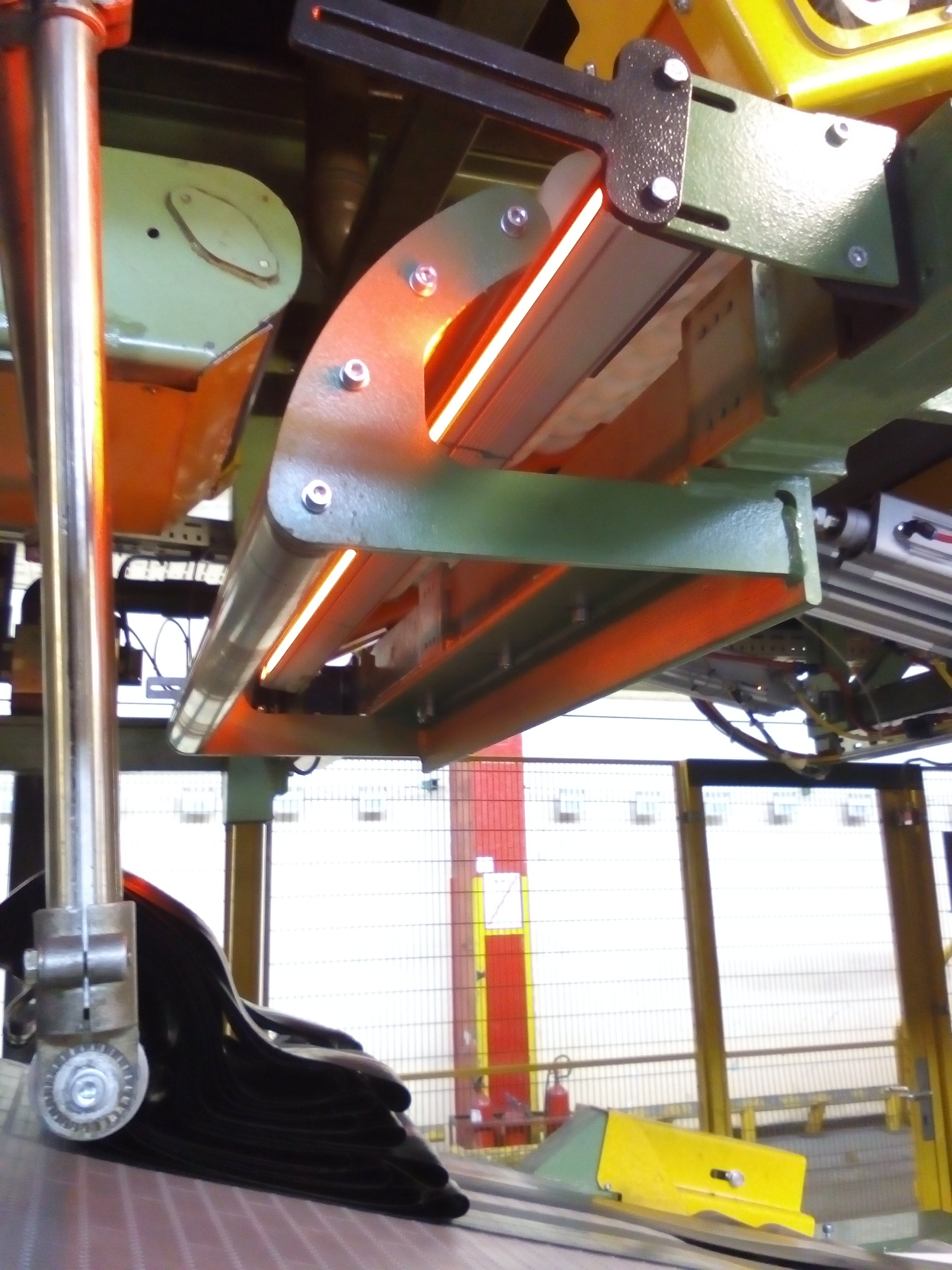 Widok z dołu na fragment konstrukcji stalowej z elementami oświetlenia LED, mocowany na śruby, z widocznymi przewodami i elementami instalacji w tle hali przemysłowej.