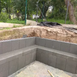 Budowa fundamentów z szarych bloczków betonowych w wykopie ziemnym, widoczne napięte sznurki wyznaczające poziom, w tle drzewa i zielona siatka ogrodzeniowa.