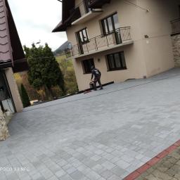 F-H-U LIB-BRUK - Nowa, szara nawierzchnia z kostki brukowej przed domem z balkonami, pracownik zamiata resztki piasku, w tle widoczne góry.