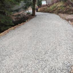 F-H-U LIB-BRUK - Świeżo utwardzona droga szutrowa w lesie, pracownik w odblaskowej kamizelce używa ubijarki w tle, drzewa po bokach, widoczna nierówność terenu.