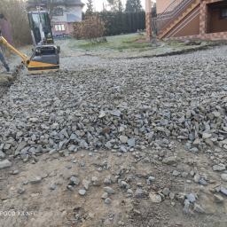 F-H-U LIB-BRUK - Pracownik obsługuje zagęszczarkę na podsypce z tłucznia przed budynkiem z brązowymi schodami, widoczny także trawnik i drzewa.