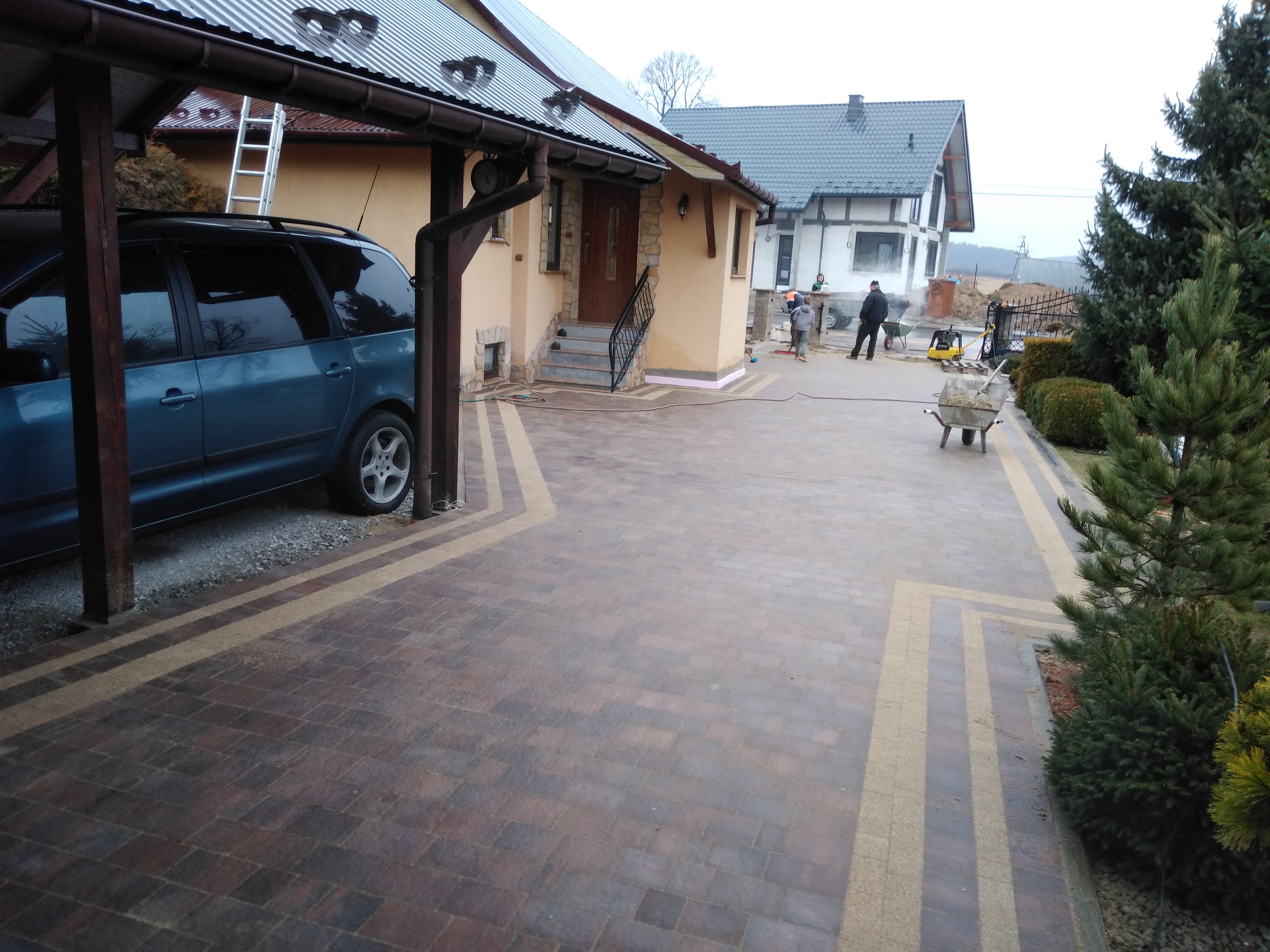 Ułożona kostka brukowa w geometryczny wzór przed domem z widocznym samochodem, pracownikami i zagęszczarką w tle.