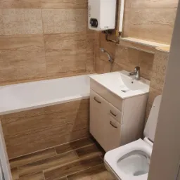 Jasna łazienka z beżowymi płytkami imitującymi trawertyn, białą wanną, szafką pod umywalkę i toaletą, oraz piecykiem gazowym nad wanną.