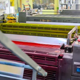 Wnętrze drukarni z rzędem wielokolorowych maszyn offsetowych, widoczne elementy sterujące i podajniki papieru, w tle pojemniki z farbami.