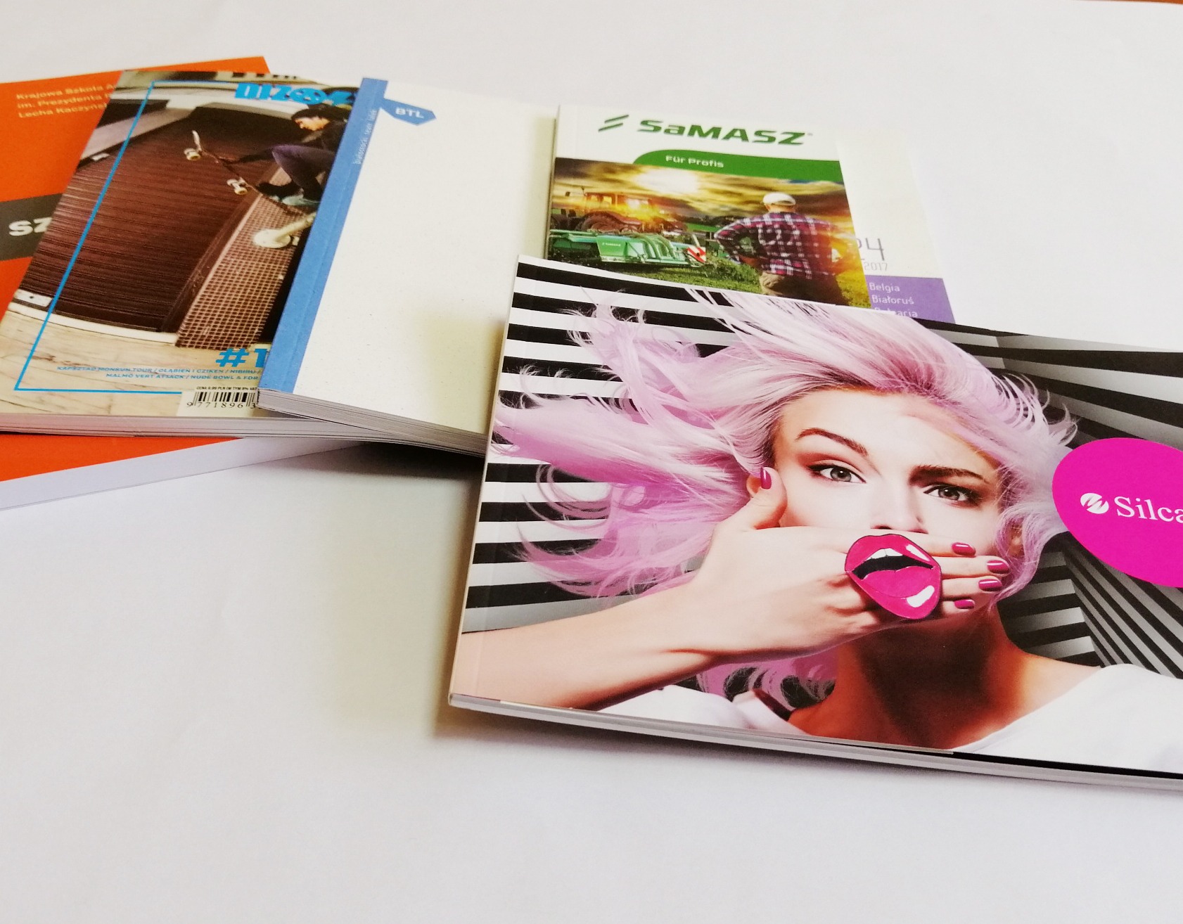 Stos różnorodnych, świeżo wydrukowanych publikacji, w tym katalogi z motywami sportowymi, rolniczymi i okładką z wizerunkiem kobiety z różowymi włosami i ustami, leżących na białym tle.