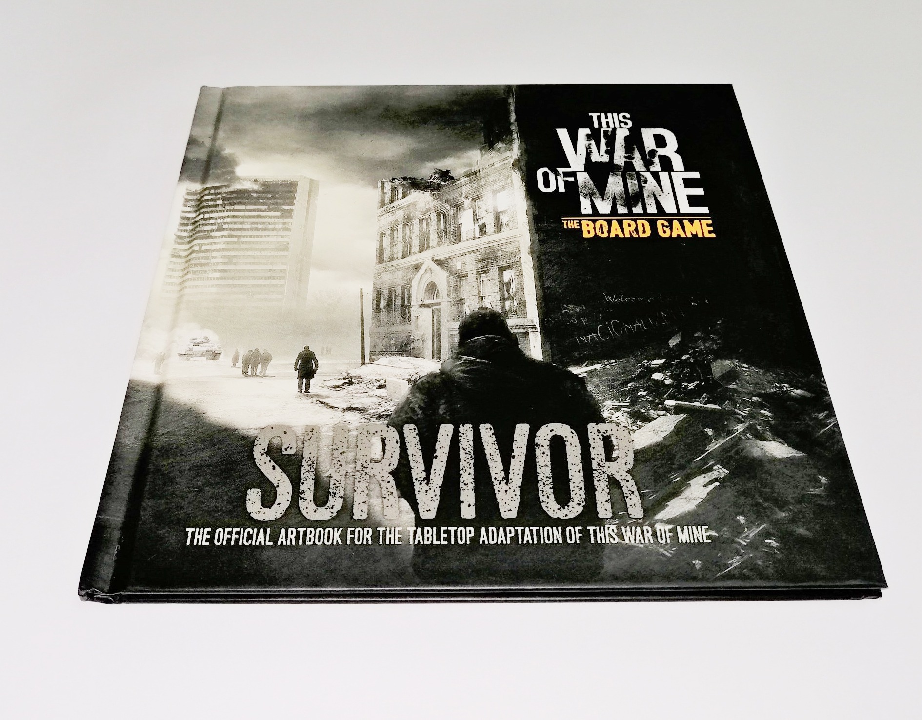 Okładka albumu z grafikami do gry planszowej 'This War of Mine: The Board Game', przedstawiająca zniszczone miasto i postać w tle.