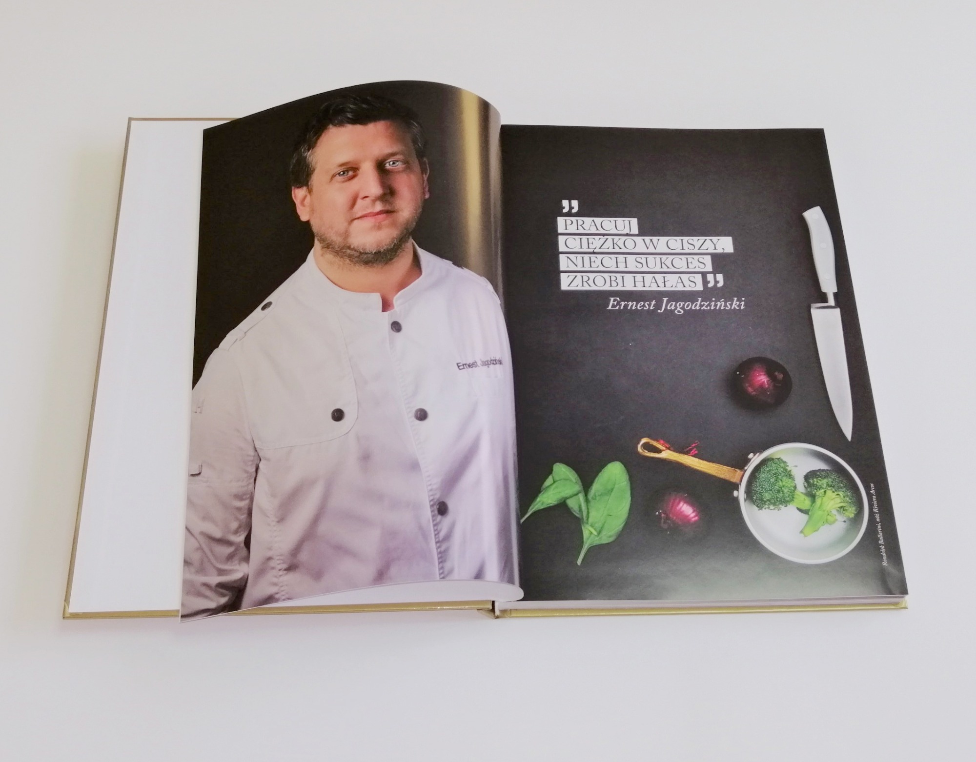 Otwarta książka z fotografią szefa kuchni w stroju roboczym i stroną z czarnym tłem, na której widnieje garnek z brokułami, cebula, szpinak i nóż oraz cytat Ernesta Jagodzińskiego.