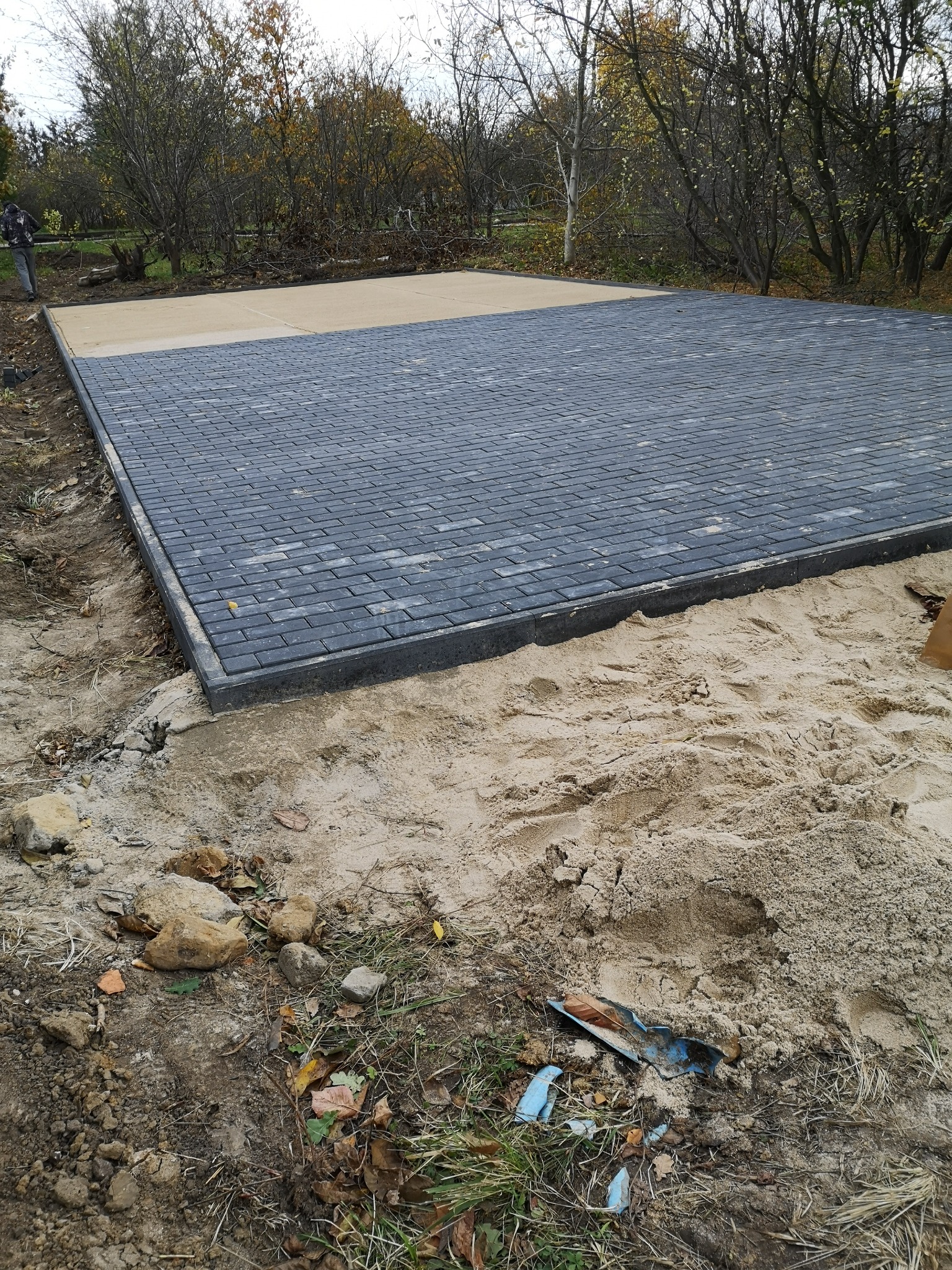 Niedokończona powierzchnia z ciemnoszarej kostki brukowej w kształcie cegiełek, obramowana szarym obrzeżem, z widoczną warstwą piasku przygotowaną pod dalszą część nawierzchni, w tle drzewa bez liści.