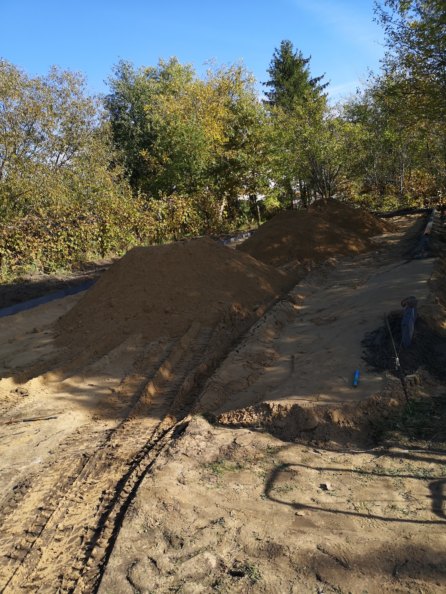 Przygotowanie terenu pod budowę: wyrównany teren z dwoma pryzmami ziemi, widoczne ślady opon, krawężniki i narzędzia w tle, błękitne niebo i drzewa w tle.