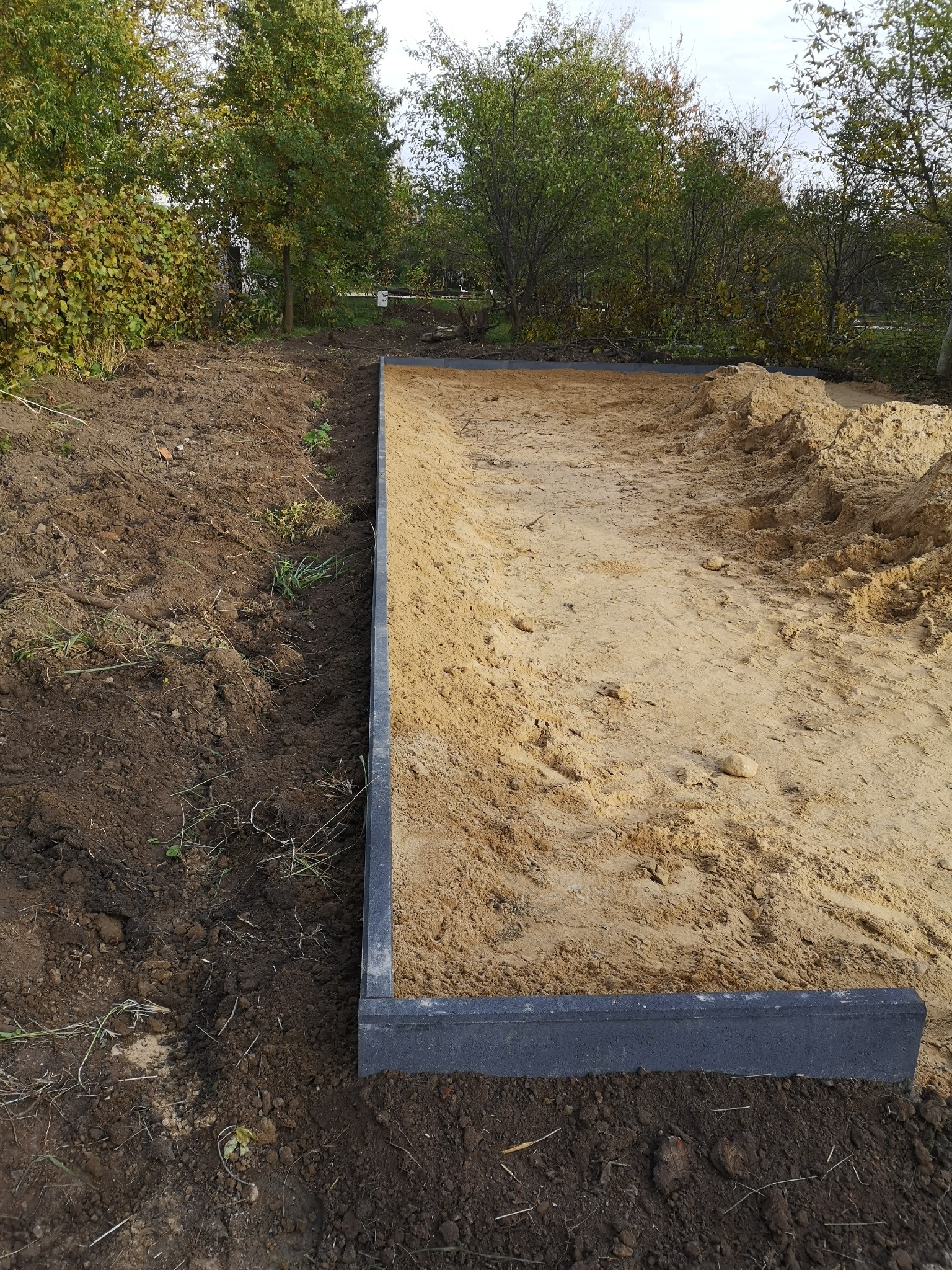 Przygotowanie podłoża pod nawierzchnię z piasku ograniczonego obrzeżem z betonowej kostki, widoczne nierówne podłoże z piasku i ziemi, w tle drzewa.