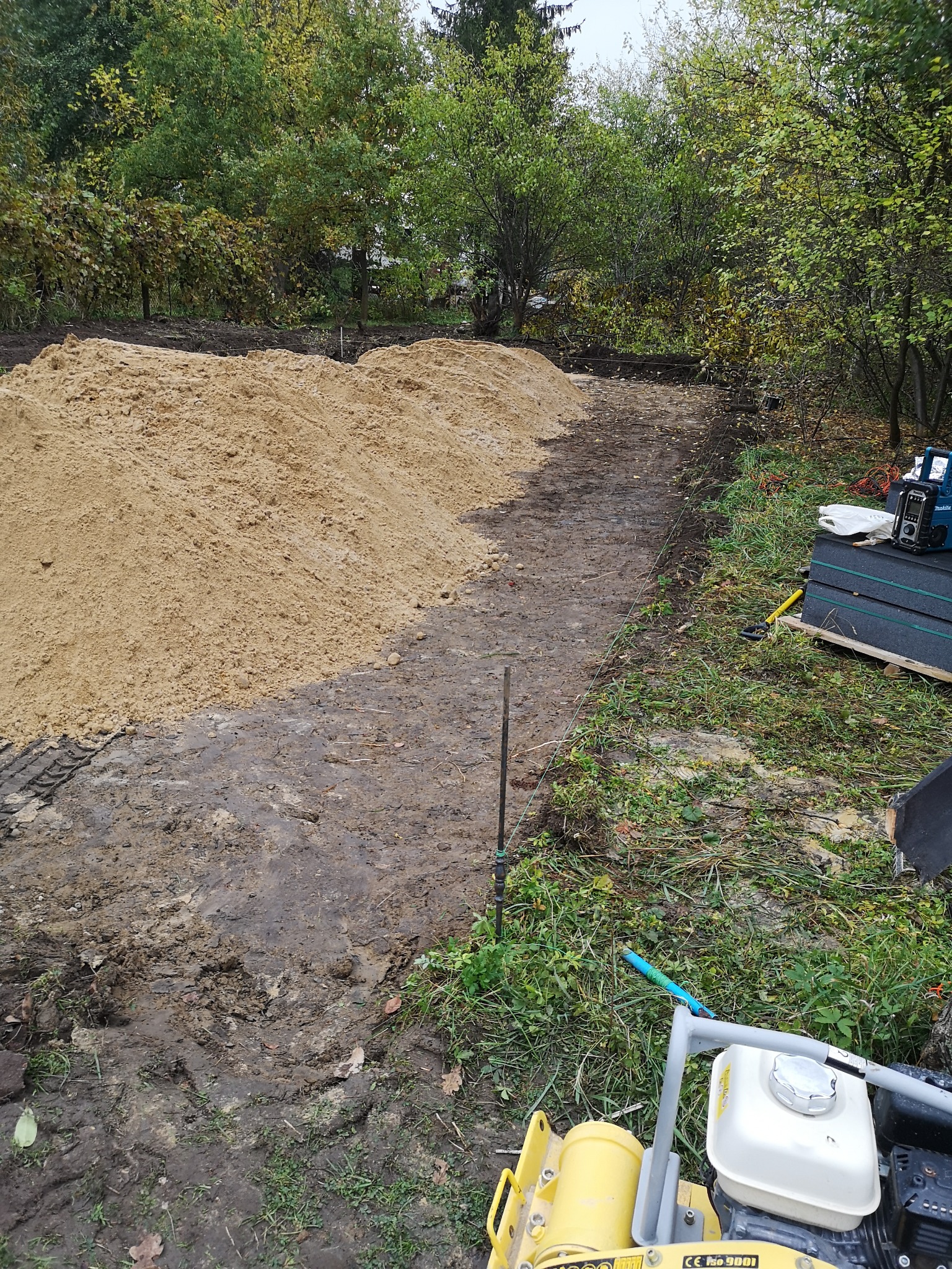 Przygotowanie terenu pod budowę: wyrównany teren z hałdą piasku, widoczne narzędzia budowlane i ubijarka w tle, zieleń drzew. Wytyczone sznurkiem granice przyszłej budowli.