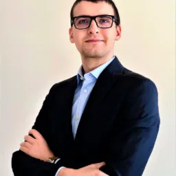 Młody mężczyzna w okularach i garniturze, z założonymi rękami, pozujący na jasnym tle.