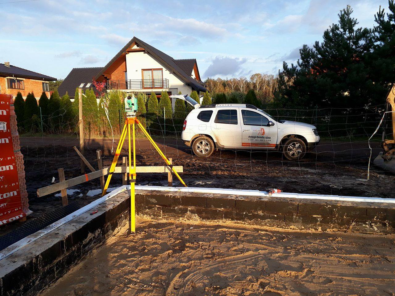 Żółty teodolit ustawiony na trójnogu, wyznaczający punkty pod budowę fundamentów domu jednorodzinnego, w tle samochód terenowy z reklamą usług geodezyjnych.