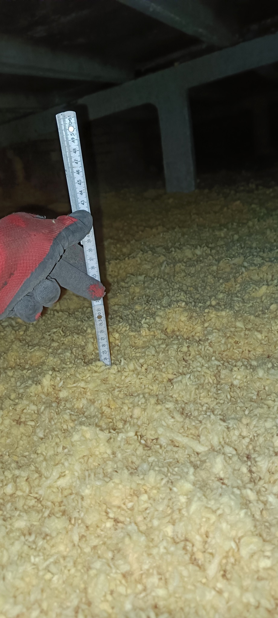 Pomiar grubości izolacji z wełny mineralnej na poddaszu, widok z bliska, ręka w roboczej rękawicy trzyma miarkę.