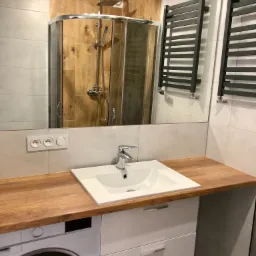 Nowoczesna łazienka z drewnianym blatem, białą umywalką, pralką w zabudowie, kabiną prysznicową z drewnianymi panelami i grzejnikami drabinkowymi.