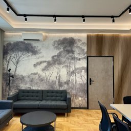 Alfa Lider - Nowoczesne wnętrze biurowe z szarą sofą, fotelem, okrągłym stolikiem kawowym i fototapetą z motywem leśnym, oświetlenie szynowe i klimatyzacja.