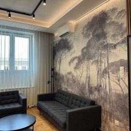 Alfa Lider - Nowoczesny salon z szarą sofą i fotelami, okrągłym stolikiem kawowym i tapetą z motywem leśnym, oświetlony listwą LED i reflektorami sufitowymi.