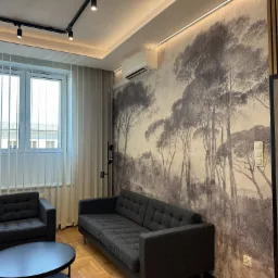 Nowoczesny salon z szarą sofą i fotelami, okrągłym stolikiem kawowym i tapetą z motywem leśnym, oświetlony listwą LED i reflektorami sufitowymi.