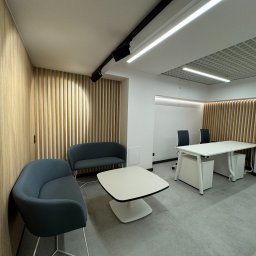 Alfa Lider - Nowoczesne wnętrze biurowe z drewnianą ścianą, szarymi sofami i białym stołem, oświetlone reflektorami sufitowymi.