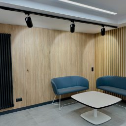 Alfa Lider - Nowoczesne wnętrze biurowe z drewnianymi panelami ściennymi, czarnym grzejnikiem żeberkowym, oświetleniem szynowym i niebieskimi sofami oraz białym stolikiem kawowym na szarej podłodze.