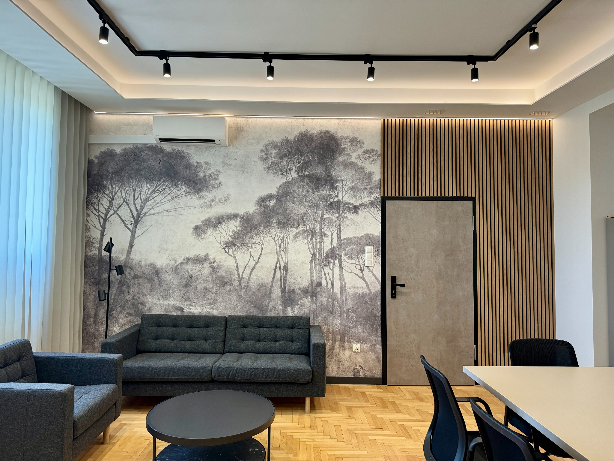 Nowoczesne wnętrze biurowe z szarą sofą, fotelem, okrągłym stolikiem kawowym i fototapetą z motywem leśnym, oświetlenie szynowe i klimatyzacja.