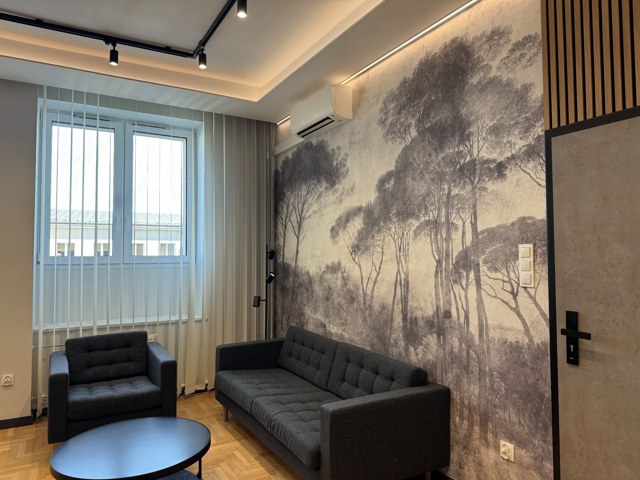 Nowoczesny salon z szarą sofą i fotelami, okrągłym stolikiem kawowym i tapetą z motywem leśnym, oświetlony listwą LED i reflektorami sufitowymi.