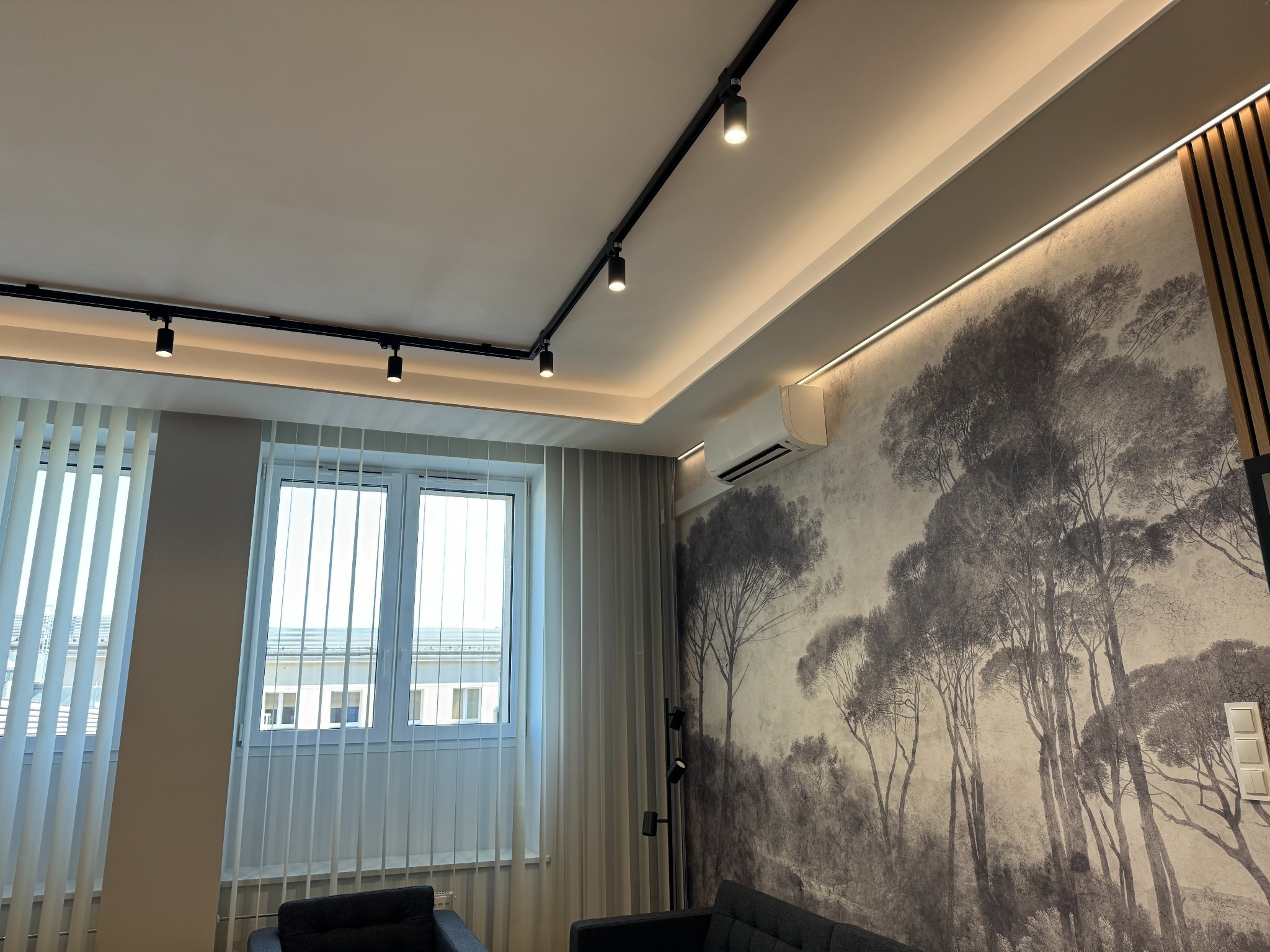 Nowoczesne oświetlenie szynowe w salonie z fototapetą leśną, widoczne również białe żaluzje pionowe i klimatyzator podsufitowy.