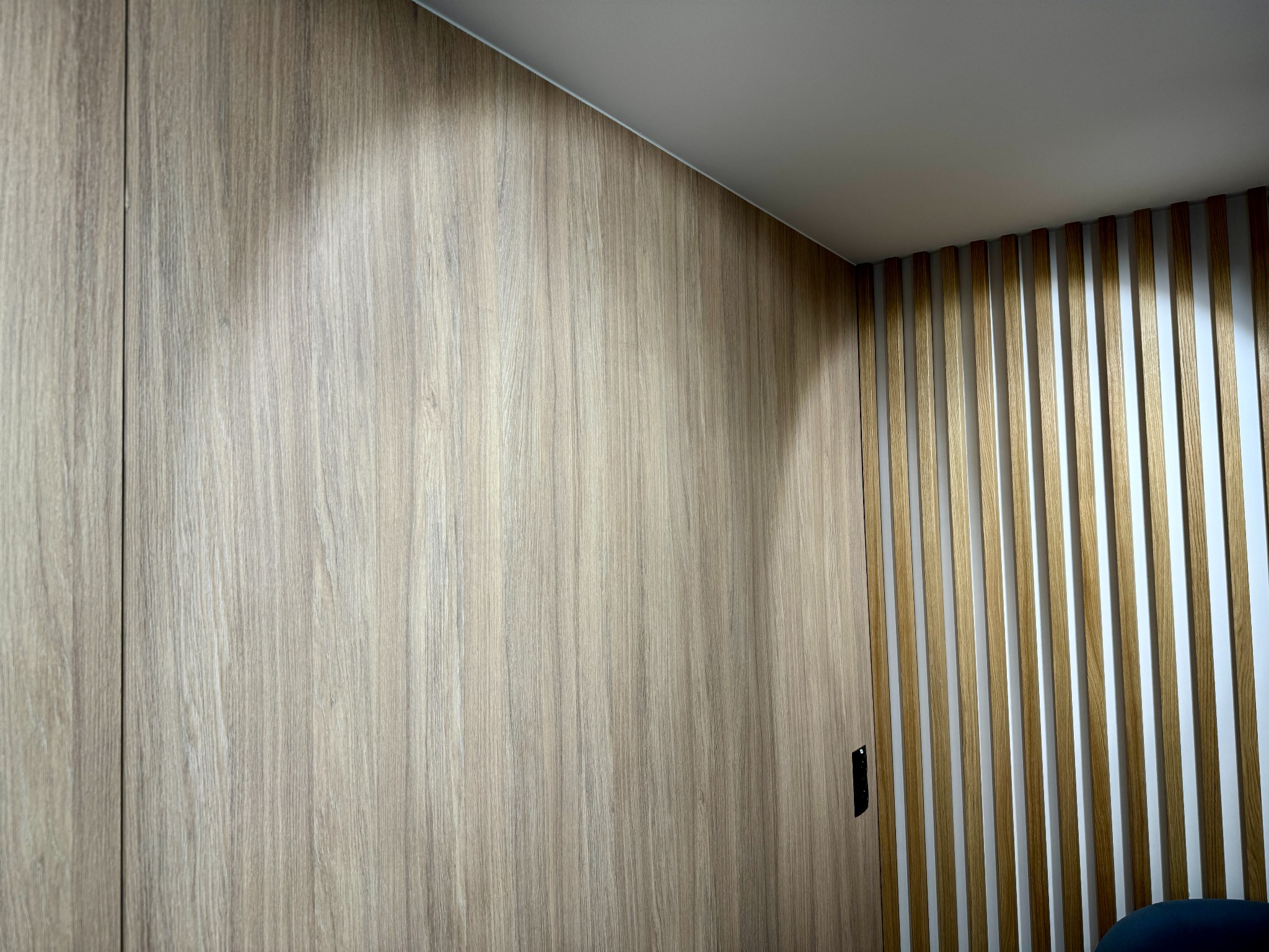 Drewniana zabudowa ściany z ukrytymi drzwiami i dekoracyjnymi lamelami pionowymi w odcieniach beżu i bieli.