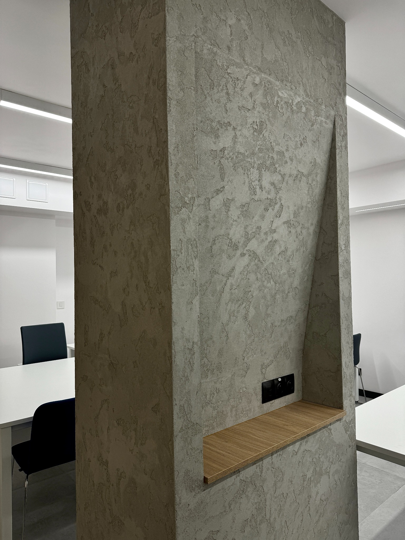 Dekoracyjny filar z wnęką i drewnianą półką w nowoczesnym biurze w Lublinie. Ściana pokryta szarą, strukturalną farbą. W tle widoczne białe biurka i czarne krzesła.