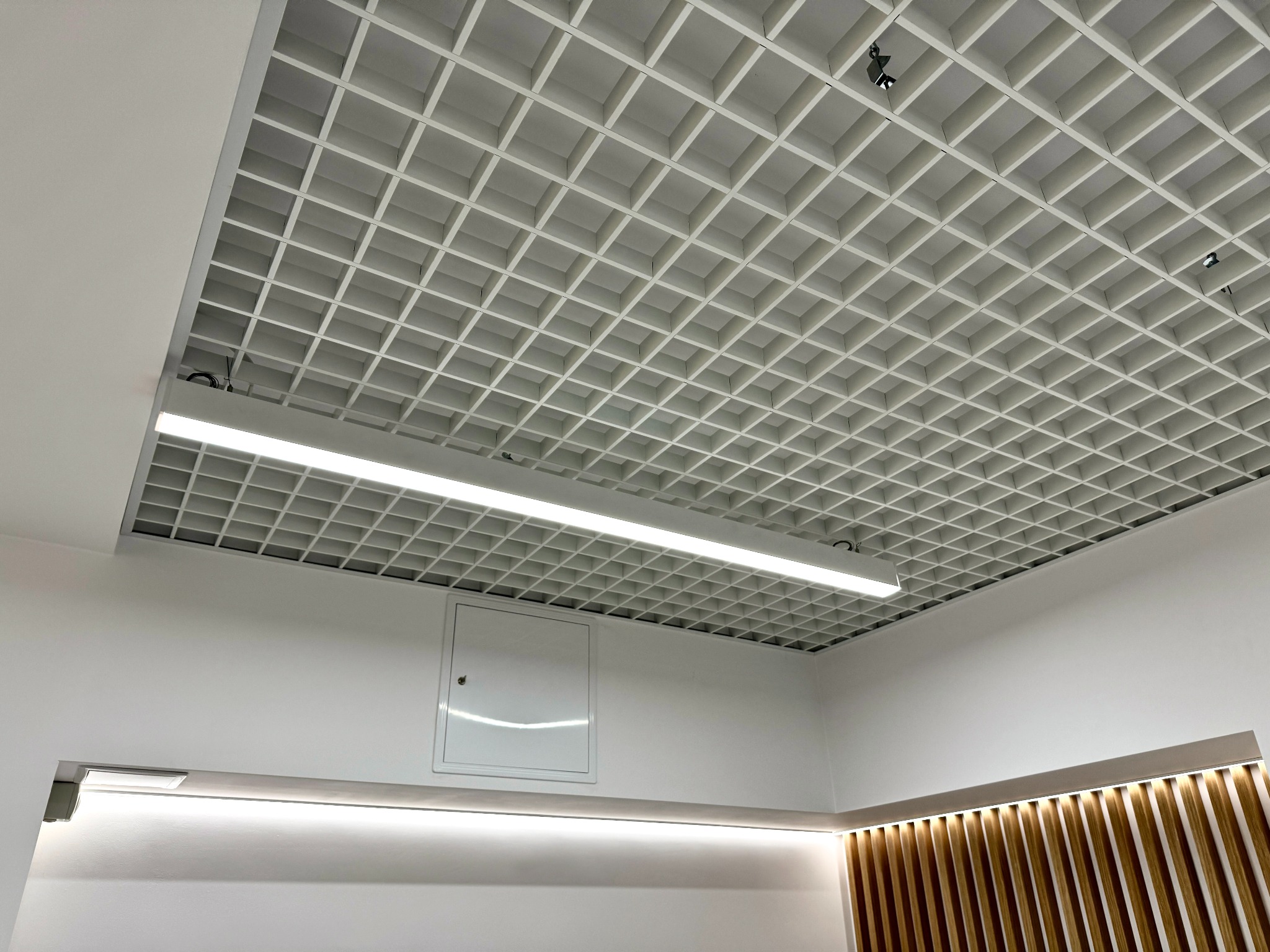 Biały sufit kasetonowy z oświetleniem liniowym LED i punktowymi reflektorami w nowoczesnym biurze, widoczny właz rewizyjny na ścianie.