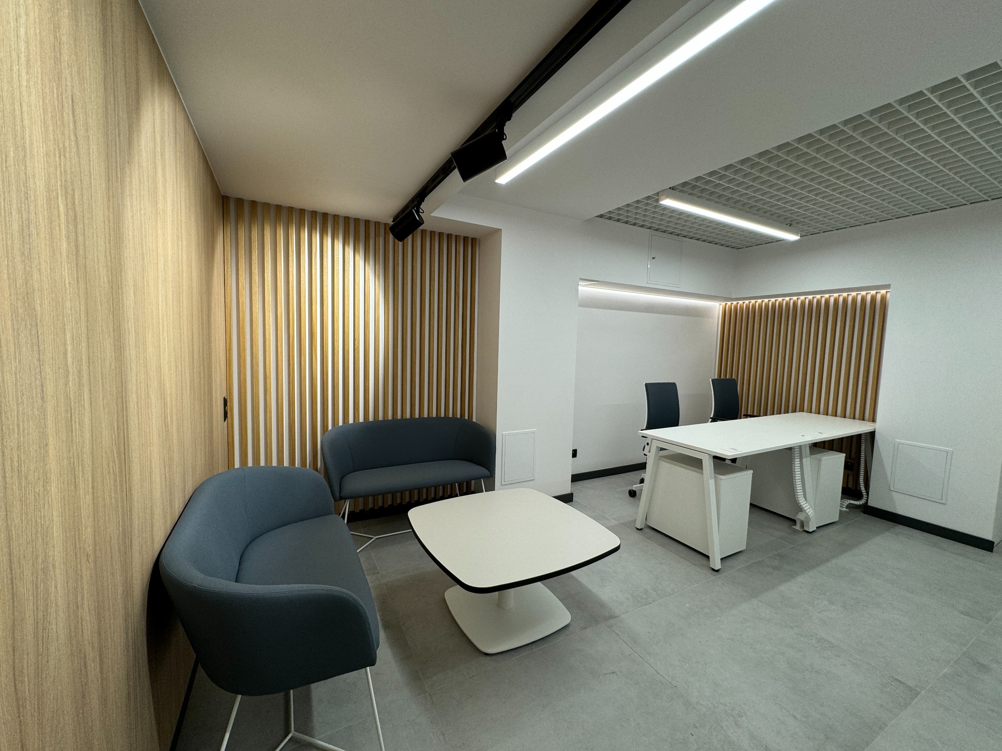 Nowoczesne wnętrze biurowe z drewnianą ścianą, szarymi sofami i białym stołem, oświetlone reflektorami sufitowymi.
