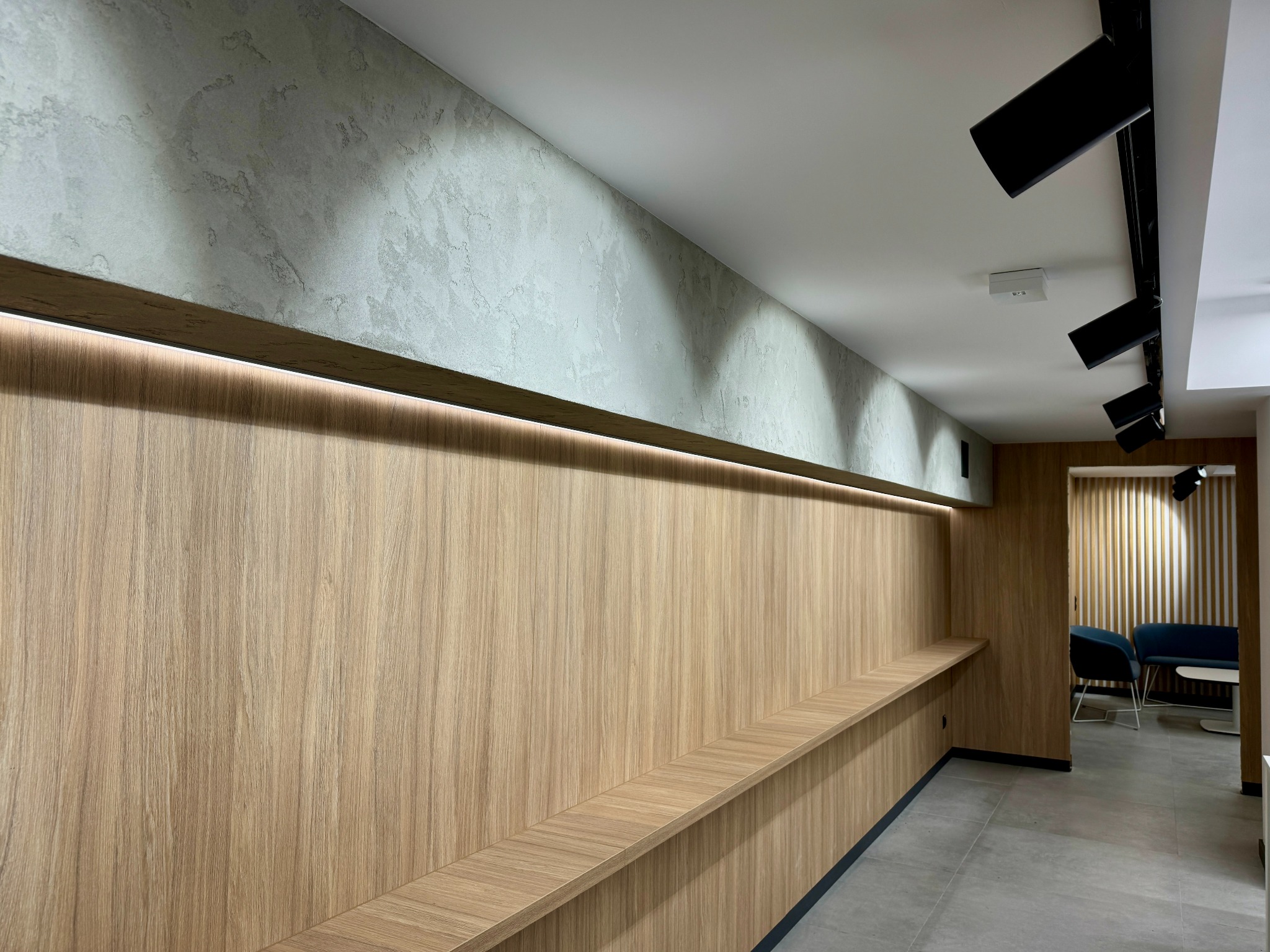 Nowoczesne wnętrze biurowe z drewnianą ścianą, podświetlaną półką i szarymi płytkami podłogowymi, oświetlone reflektorami sufitowymi.