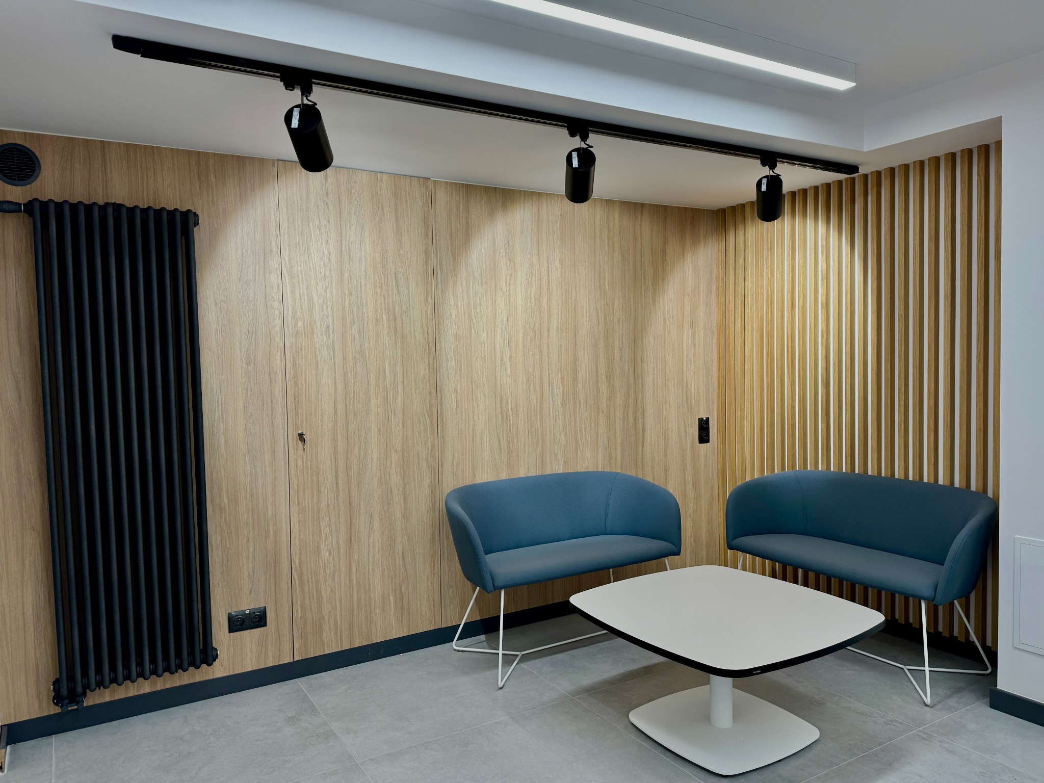 Nowoczesne wnętrze biurowe z drewnianymi panelami ściennymi, czarnym grzejnikiem żeberkowym, oświetleniem szynowym i niebieskimi sofami oraz białym stolikiem kawowym na szarej podłodze.