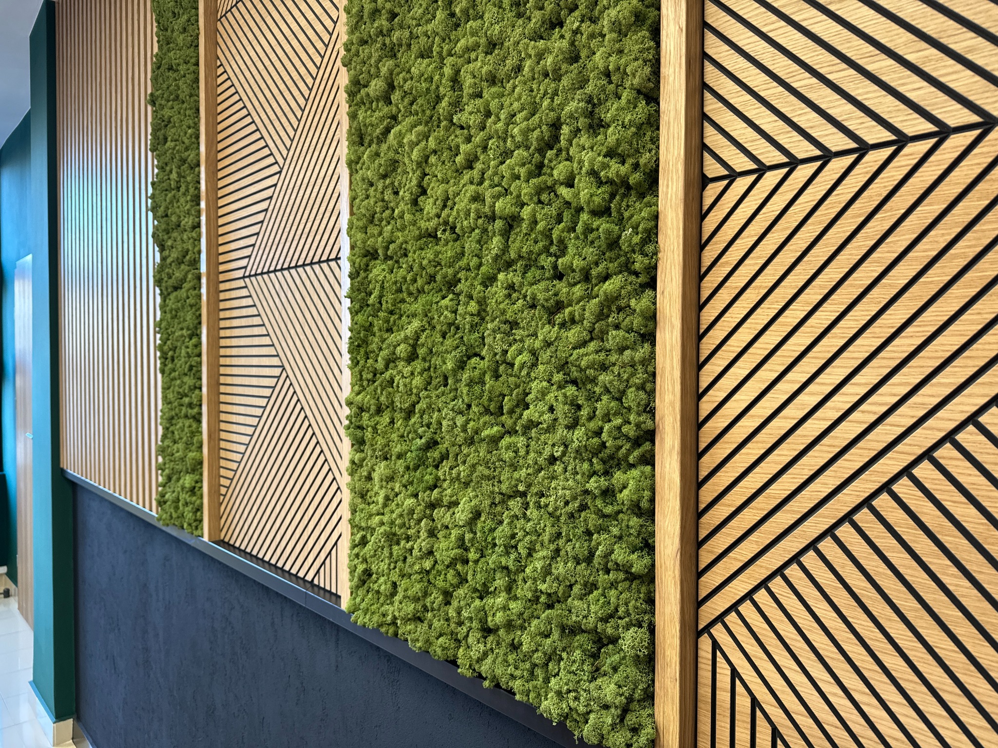 Fragment ściany z dekoracyjnymi panelami: drewniane listwy pionowe, panel z mchu chrobotka reniferowego oraz panel z ukośnymi listwami tworzącymi geometryczny wzór, całość oddzielona czarną listwą.