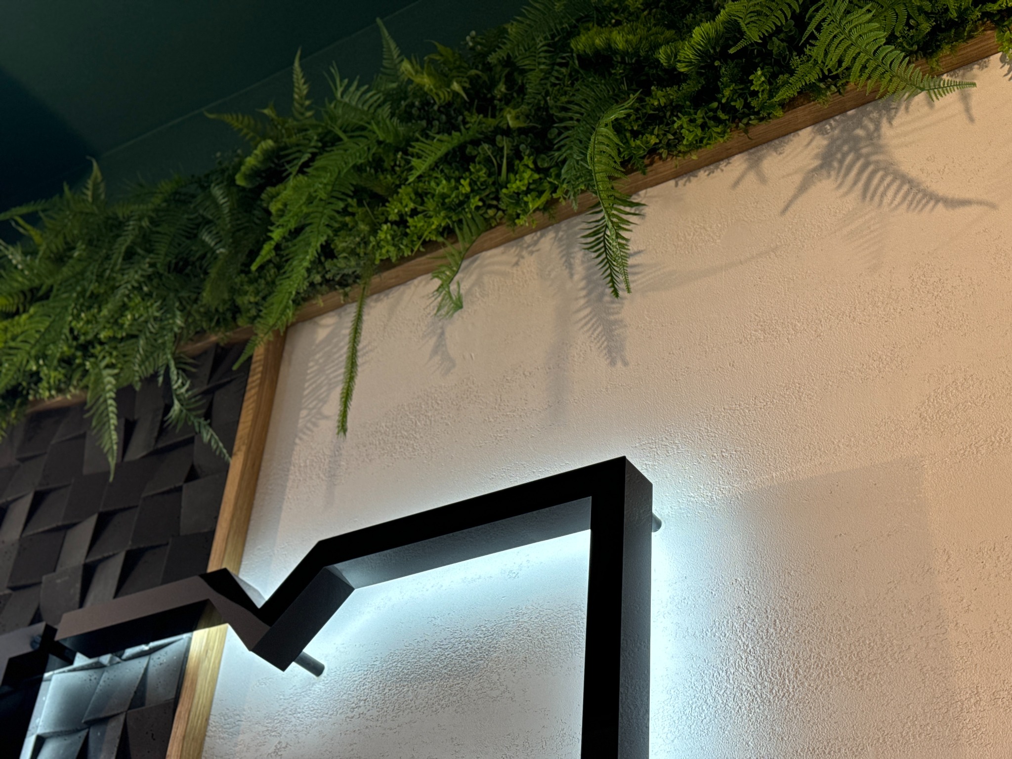 Ściana z dekoracyjnym panelem 3D, roślinami w drewnianej ramie i podświetlanym, geometrycznym elementem dekoracyjnym.