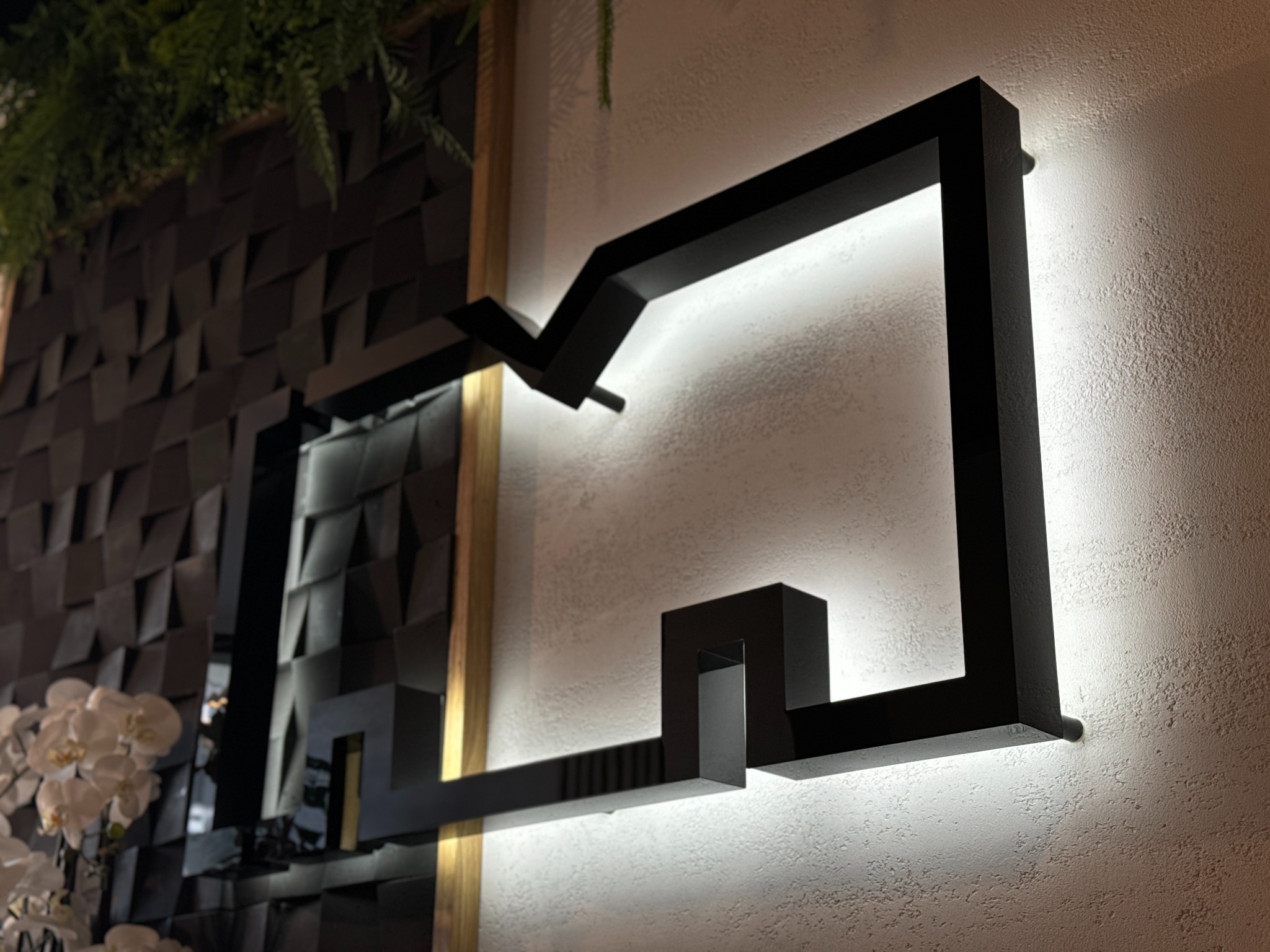 Czarna, podświetlana konstrukcja geometryczna na teksturowanej ścianie, obok dekoracyjna ściana z trójwymiarowych paneli i rośliny.