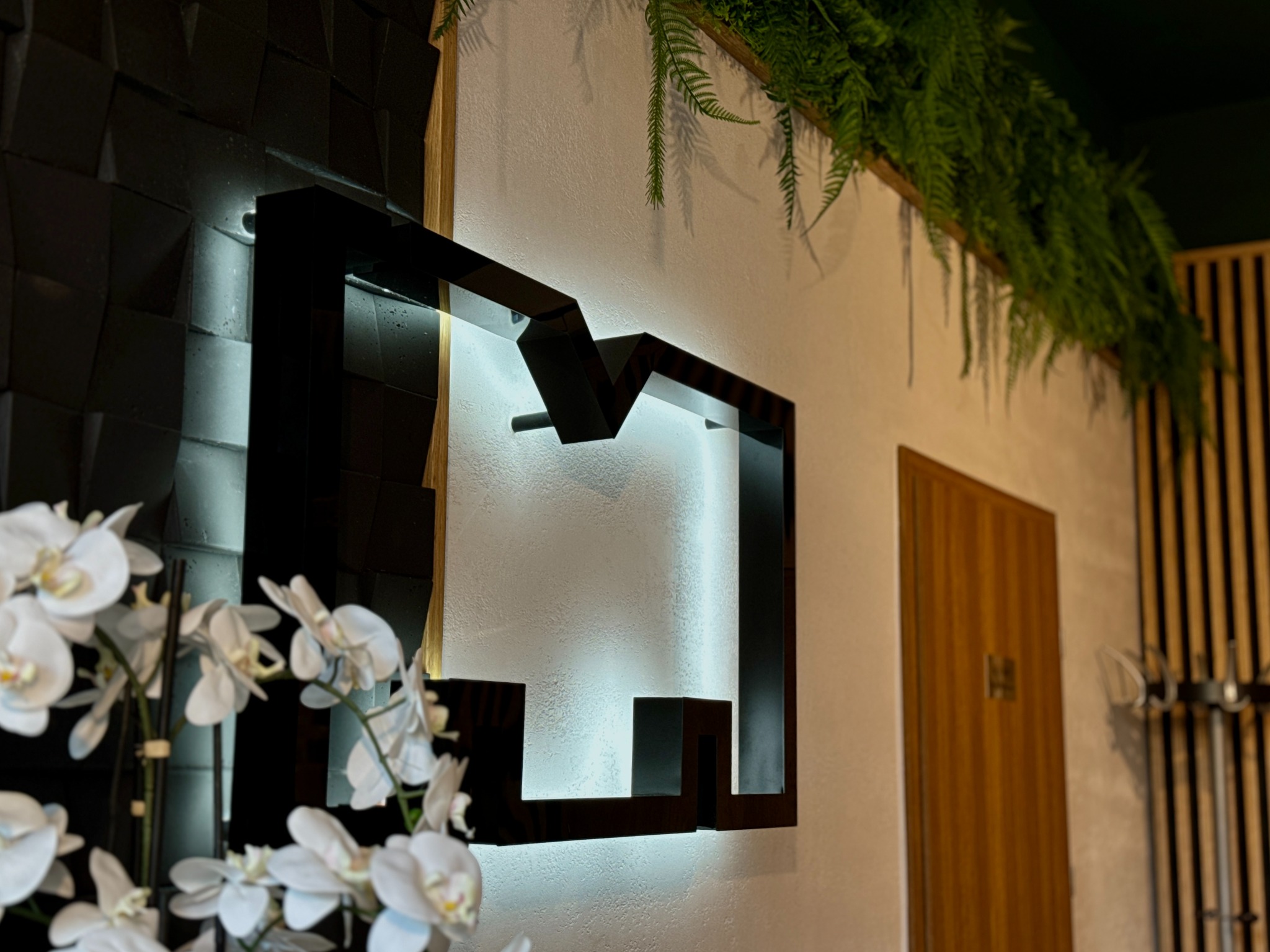Dekoracyjna, podświetlana konstrukcja na ścianie w Lublinie, obok czarnych, trójwymiarowych paneli i białych orchidei, z elementami zieleni zwisającej z góry.