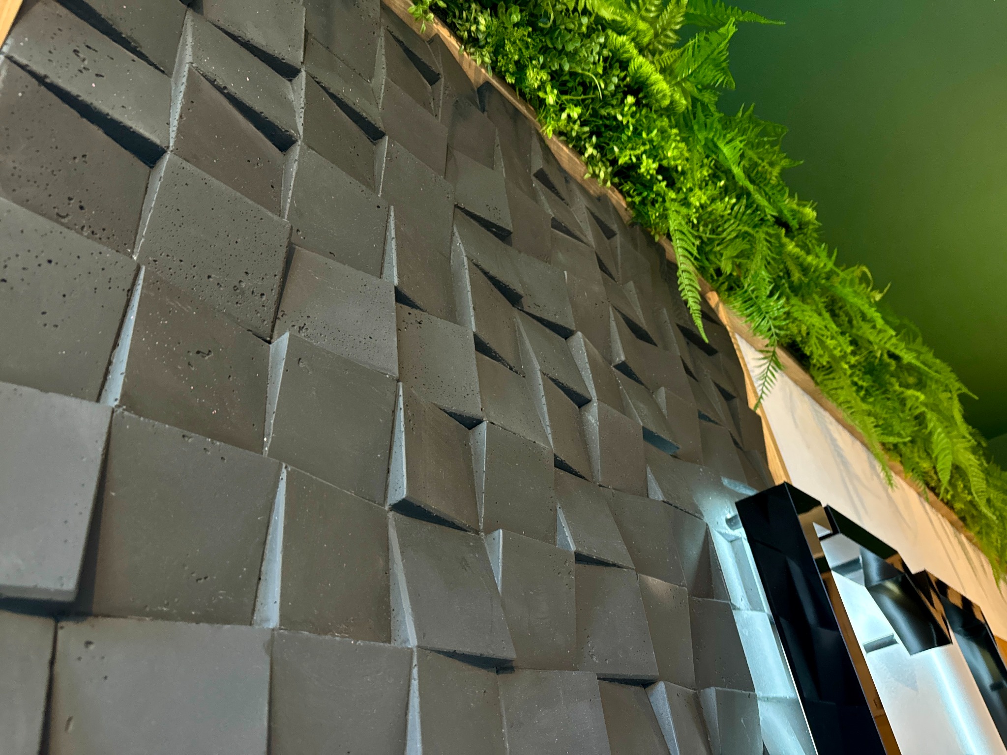 Dekoracyjna ściana z trójwymiarowych, betonowych paneli w odcieniach szarości, u góry ozdobiona zielonymi roślinami i paprociami, oświetlona punktowym światłem.