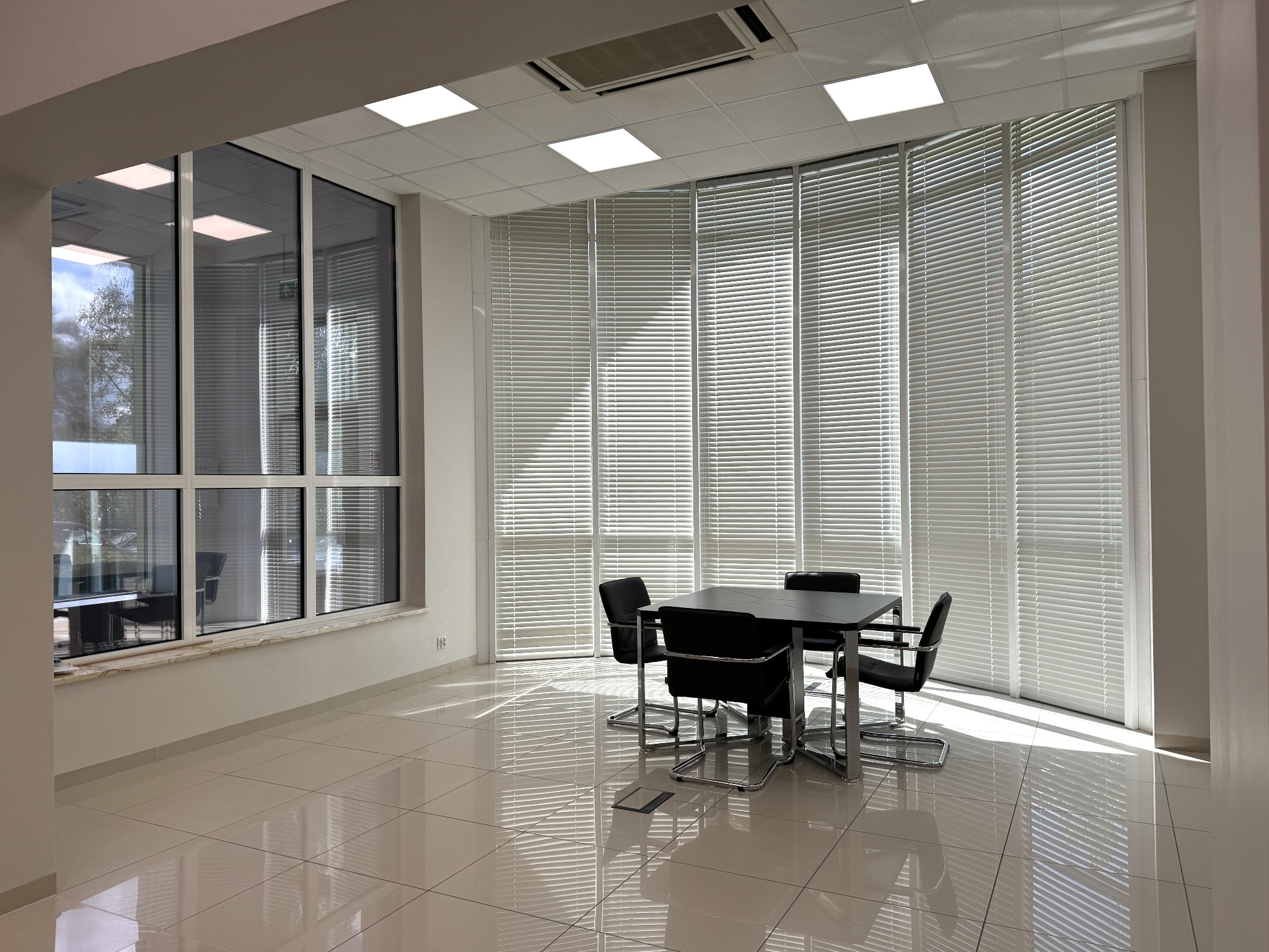 Nowoczesne biuro z dużymi oknami z żaluzjami, czarnym stołem konferencyjnym i krzesłami na chromowanych nogach, jasna podłoga z połyskiem, klimatyzacja sufitowa.