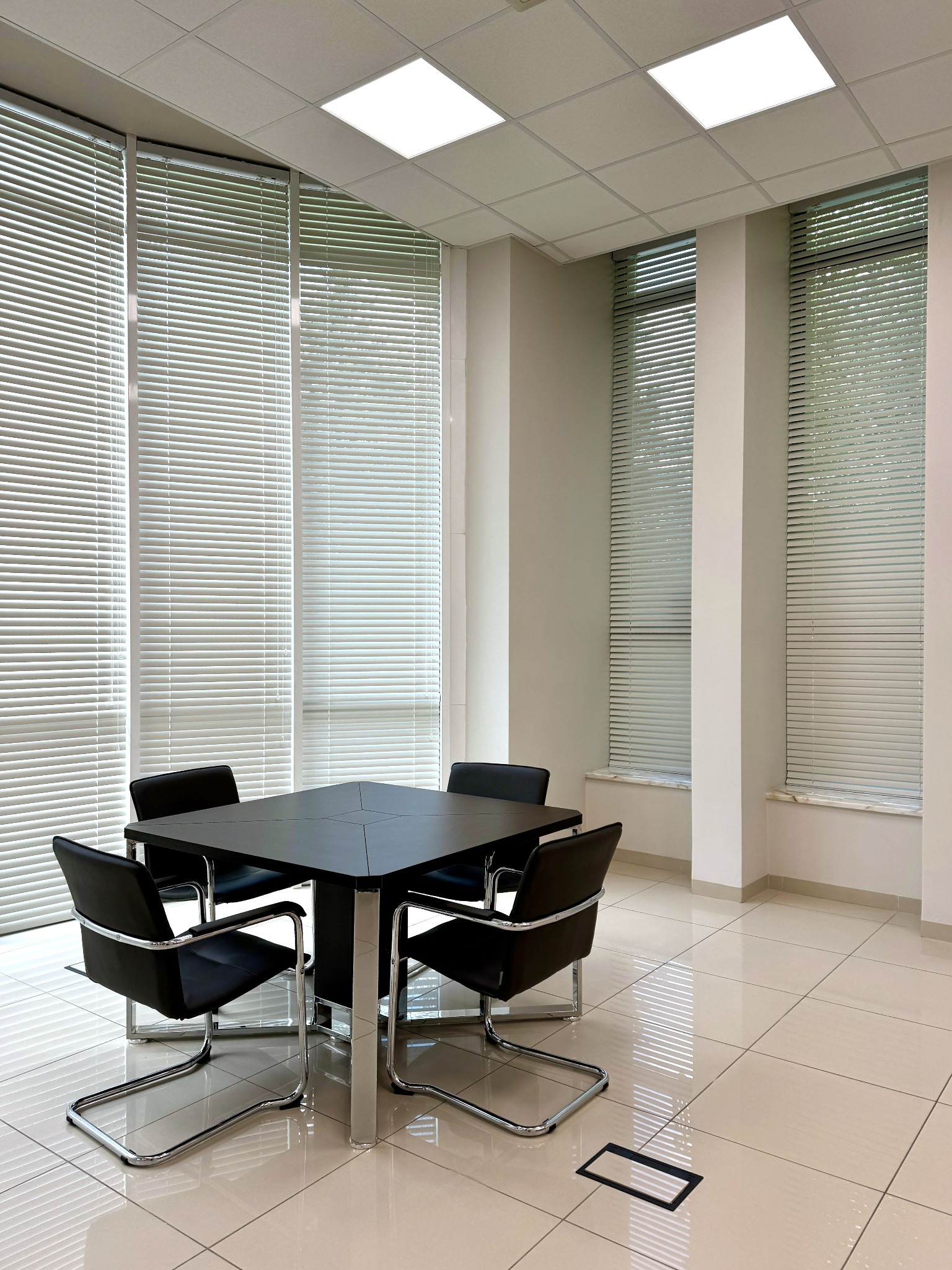 Nowoczesna sala konferencyjna z kwadratowym, czarnym stołem i czterema skórzanymi krzesłami na chromowanych nogach, oświetlona jasnym światłem z paneli sufitowych i naturalnym światłem wpadającym...