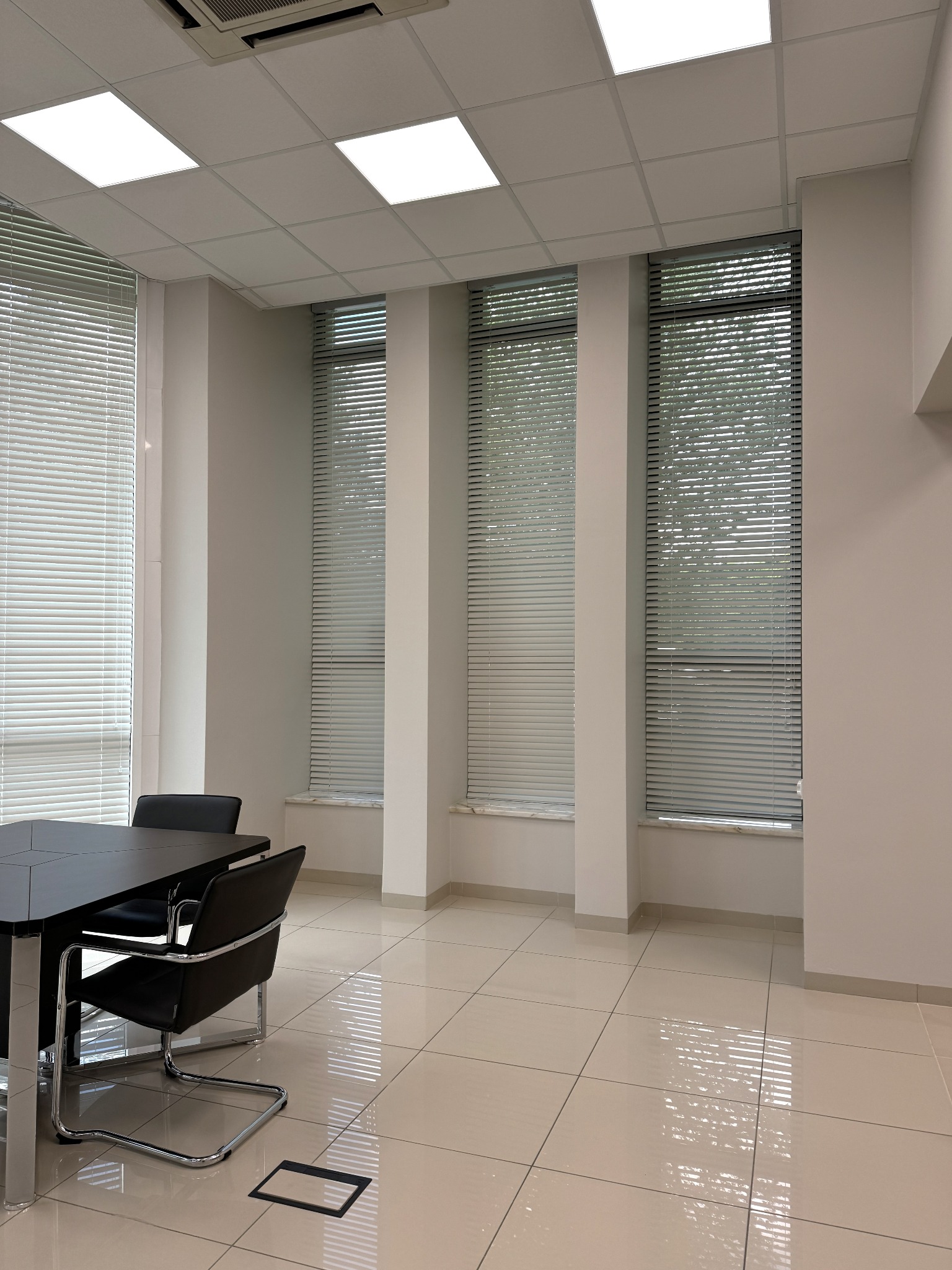 Jasne, minimalistyczne wnętrze biura z dużymi oknami z żaluzjami, czarnym stołem konferencyjnym i krzesłami na chromowanych nogach, błyszcząca podłoga z płytek.