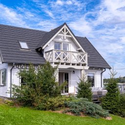 Bobieni Fotografia Wiesław Bobieni - Biały dom z szarą dachówką i balkonem z drewnianą balustradą, otoczony zielenią. Widok z przodu, słoneczny dzień, fragment schodów wejściowych z boku.